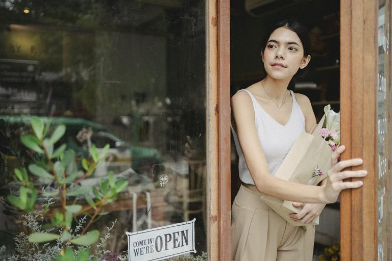 Young female in trendy outfit standing near entrance of modern flower shop with flowers in hand