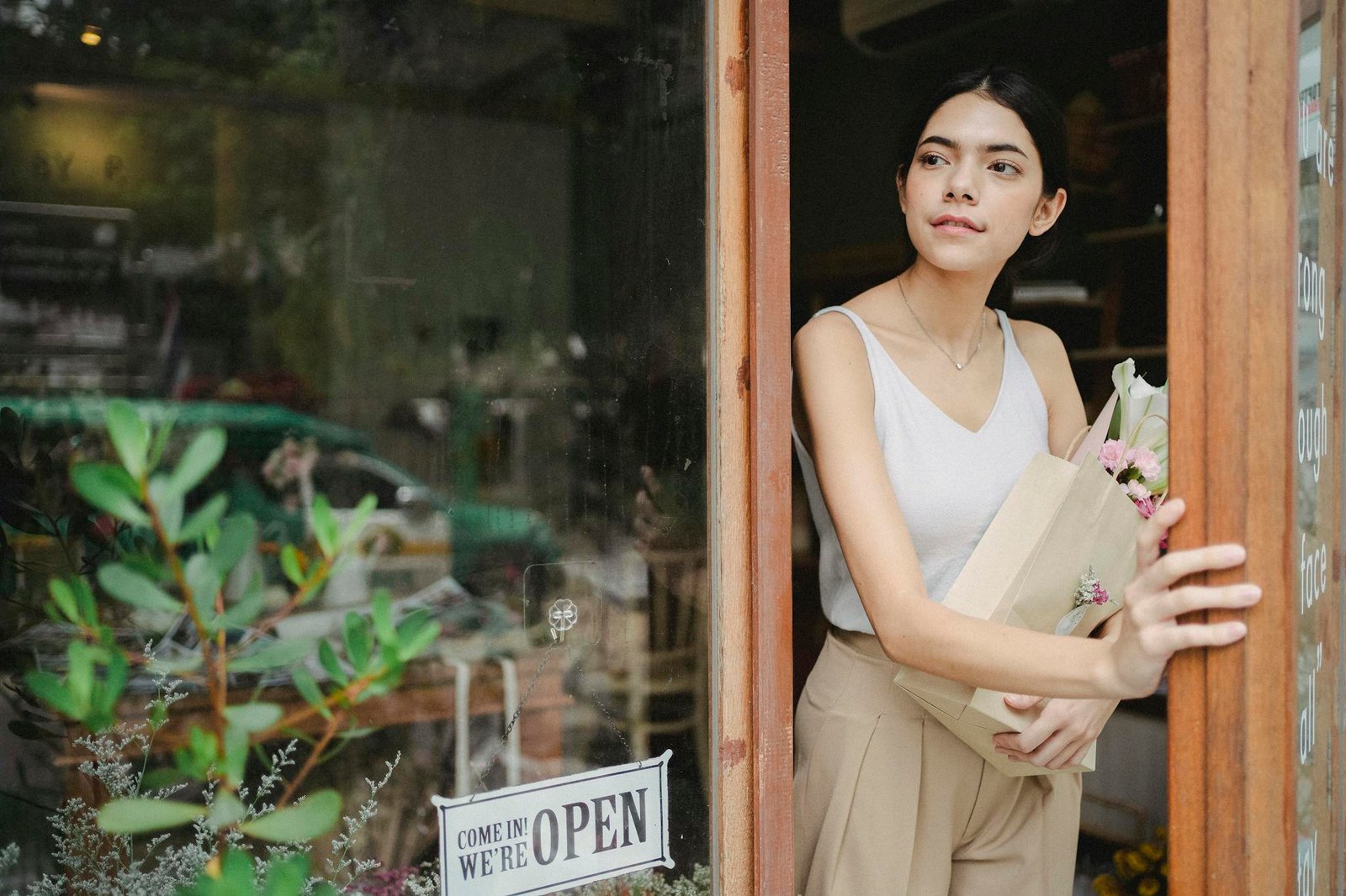 Young female in trendy outfit standing near entrance of modern flower shop with flowers in hand